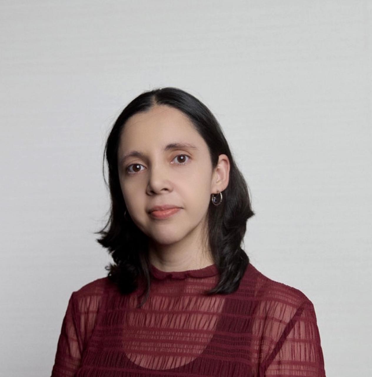 A person with shoulder-length dark hair wearing a maroon, semi-transparent top and small hoop earrings is facing the camera against a plain light background.