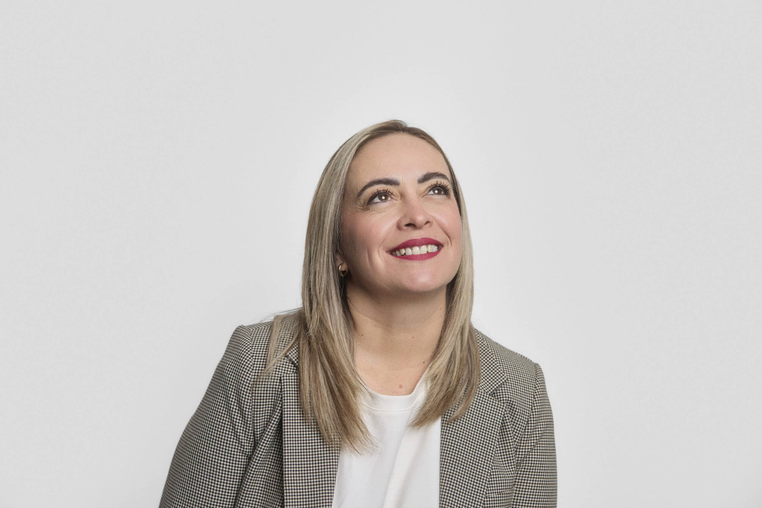 A woman with straight blonde hair, wearing a gray checked blazer and white top, looks up and smiles against a plain white background.