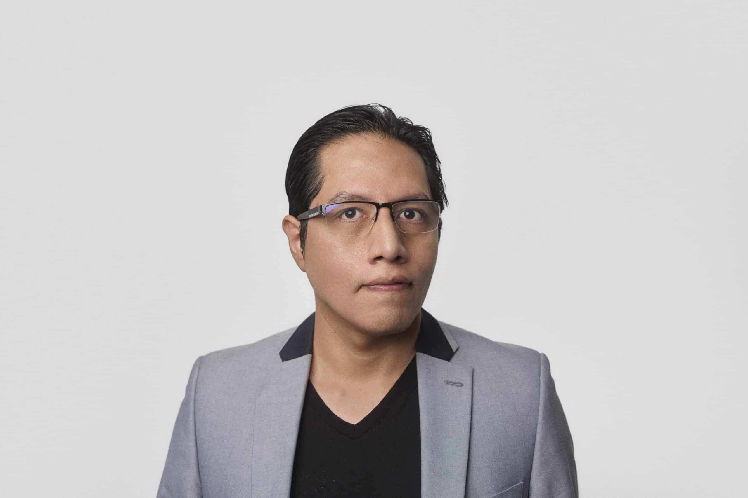 A person with short dark hair and glasses is wearing a light gray blazer over a black shirt, standing against a plain white background and looking slightly to the side.