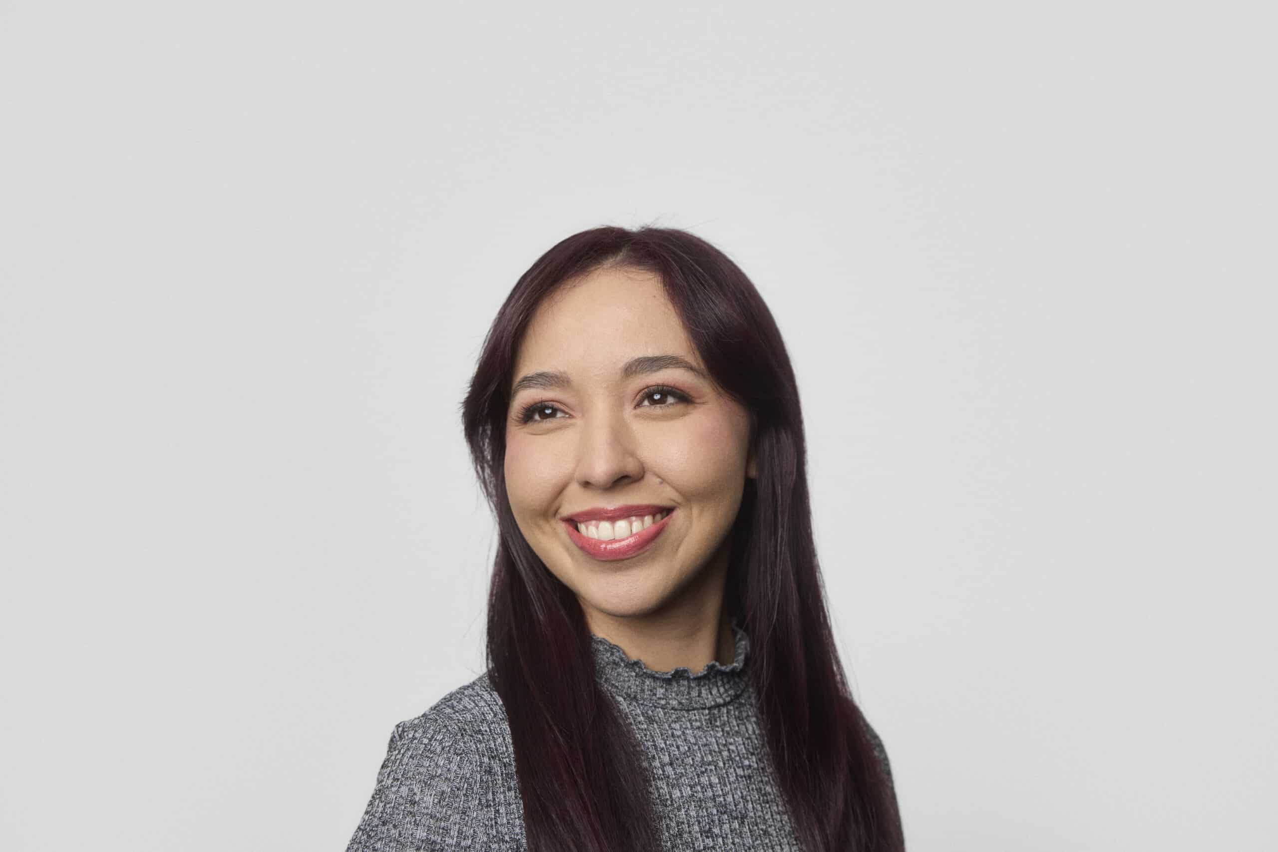 A young woman with long, dark brown hair and a gray, high-neck sweater is smiling and looking slightly to the side against a plain light gray background.