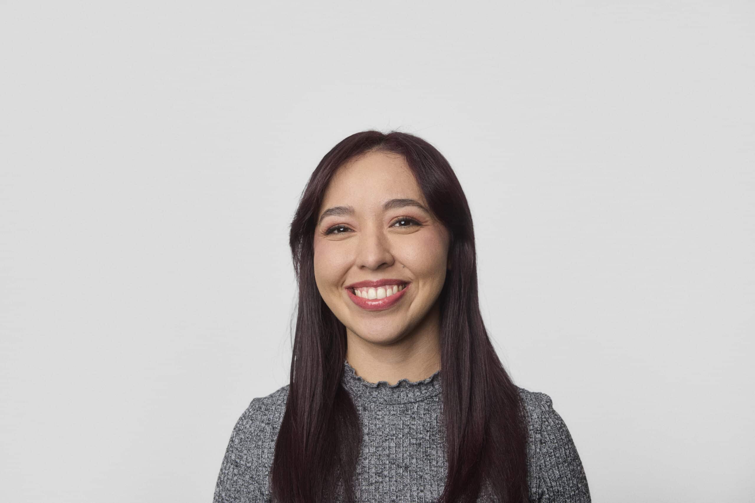 A young woman with long, straight, dark hair is smiling and looking at the camera. She is wearing a gray textured top and standing against a plain light gray background.