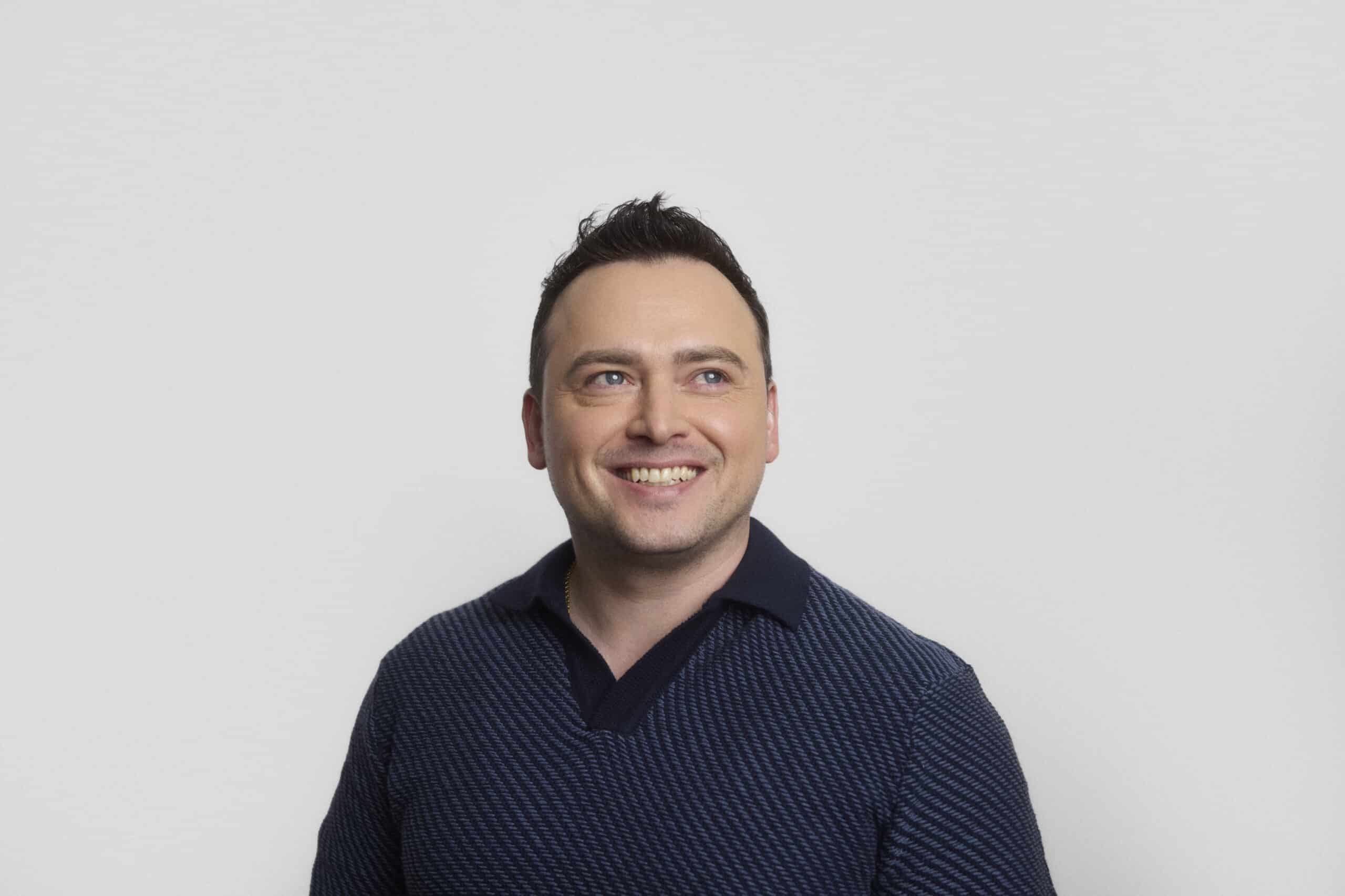 A man with short dark hair is smiling and looking slightly to his left. He is wearing a dark blue collared shirt with a textured pattern. The background is plain and light-colored.