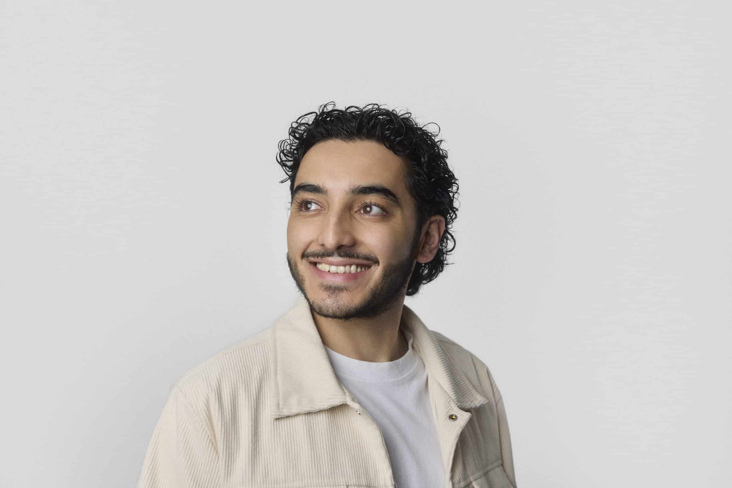 A man with curly dark hair and a beard is smiling while looking to his left. He is wearing a light-colored jacket over a white shirt. The background is plain and light gray.