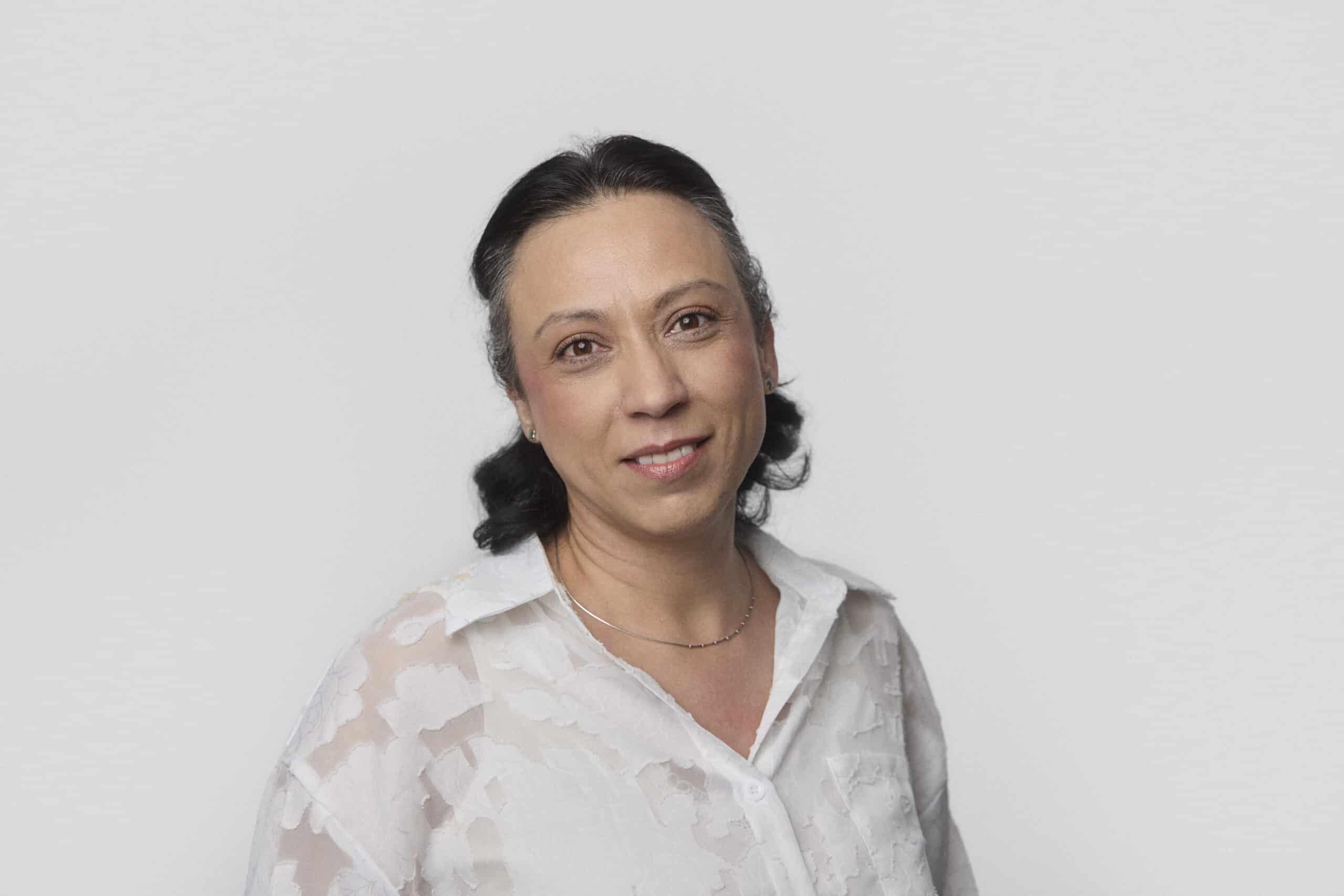 A woman with dark hair pulled back is wearing a white patterned blouse and a thin necklace, standing against a plain light gray background. She is looking at the camera with a slight smile.