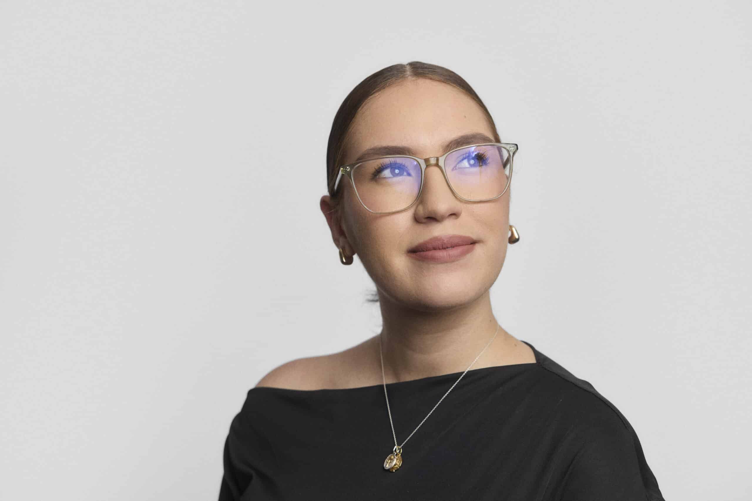 A woman with brown hair tied back wears glasses, gold earrings, a necklace, and a black off-the-shoulder top. She looks slightly upward and to the side, smiling softly against a plain light background.