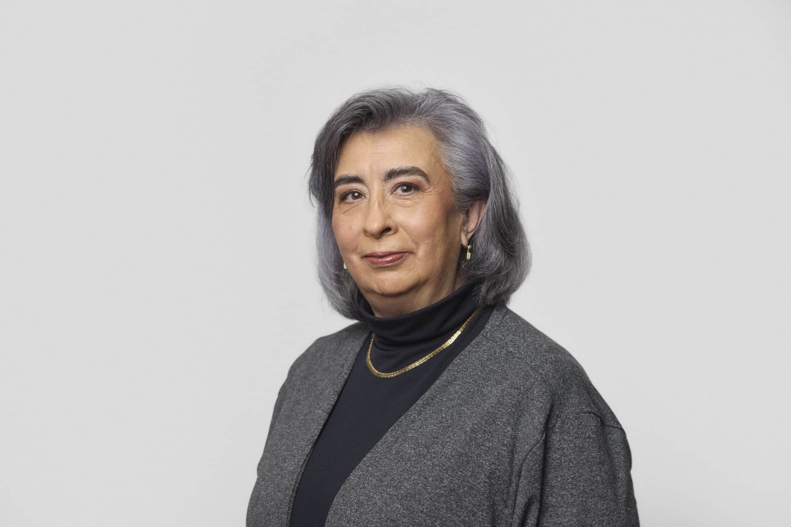 An older woman with gray hair wearing a dark turtleneck, a gray cardigan, a gold necklace, and gold hoop earrings stands against a plain light gray background, looking at the camera.