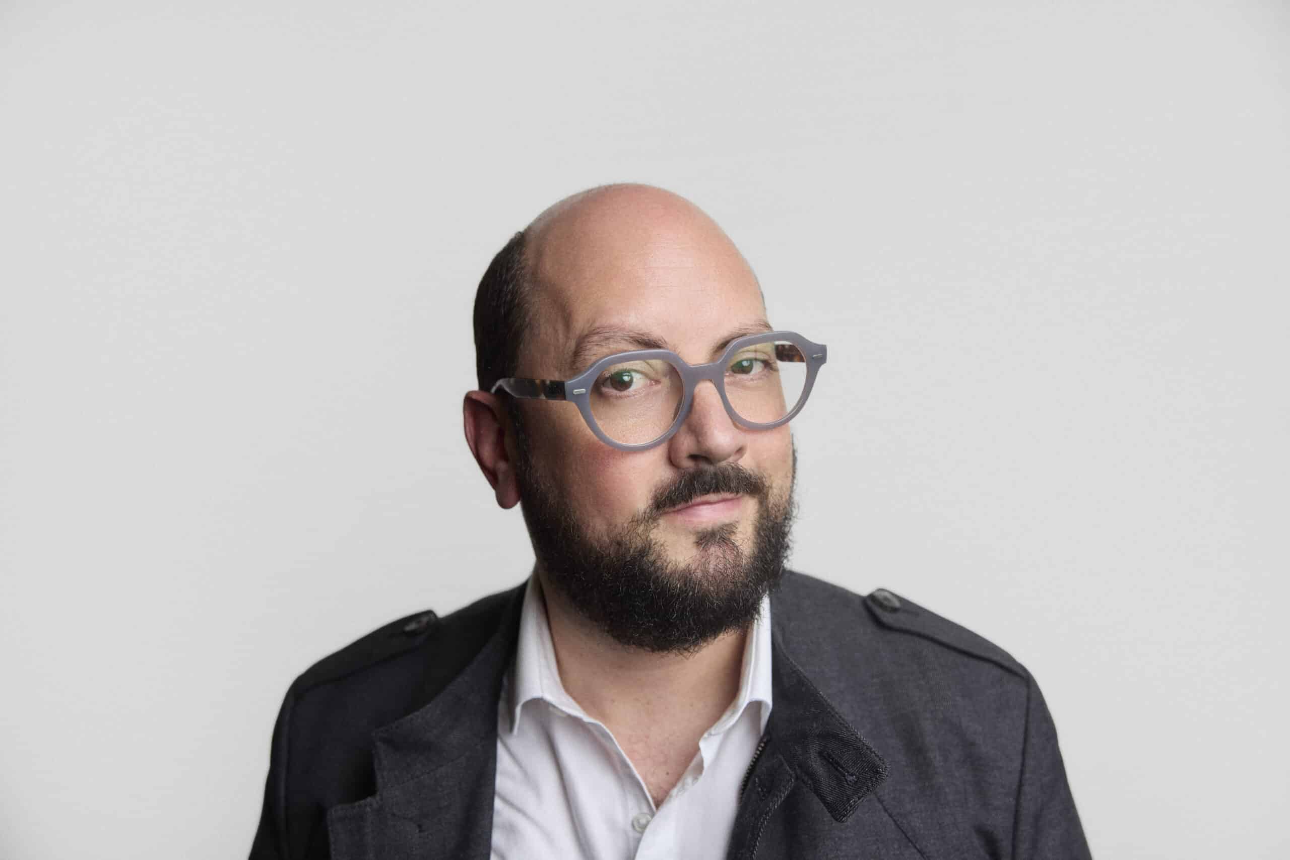 A man with a bald head and dark beard, wearing round gray glasses and a dark jacket over a white collared shirt, poses against a plain light gray background.