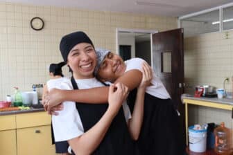 Two people wearing aprons and head coverings are standing in a kitchen, smiling and hugging each other. A third person is visible in the background. The kitchen has yellow cabinets and tiled walls.