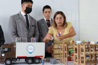 A woman in a yellow striped shirt arranges toy figures and structures on a table while two young men in gray suits observe. A toy truck labeled Jóvenes con Entrega is also present on the table.