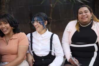 Three people sit side by side outdoors, smiling and laughing. One wears a peach top, another wears glasses and a white shirt with black overalls, and the third wears a black and white dress with a light pink jacket.