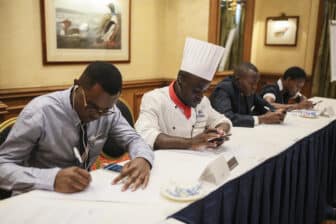 Four people sit at a table, two writing on paper and two using their phones. One person is wearing a chef’s hat and uniform. The setting appears to be a formal meeting or judging event in a decorated room.