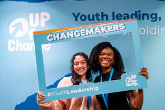 Two women smile while holding a blue frame labeled “CHANGEMAKERS” and “#YouthLeadership” at an event with “UP for Change” and “Youth leading” signs in the background.