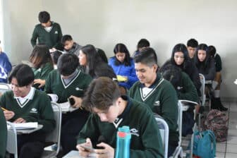 A classroom of high school students in green uniforms sit at desks, working on assignments or looking at their phones. Some students are writing, and others are reading or talking. The room has white walls and tiled flooring.