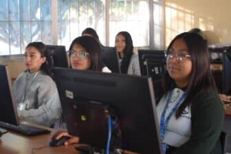 Five young women sit at computer desks in a classroom, facing forward and looking at their monitors. The room has large windows with bars, letting in natural light. The atmosphere appears focused and studious.