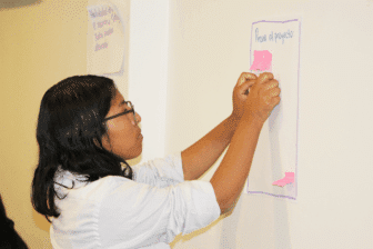 A person with long dark hair and glasses attaches pink sticky notes to a large sheet of paper taped to a wall. The paper has text at the top. Another sheet with writing is partially visible in the background.