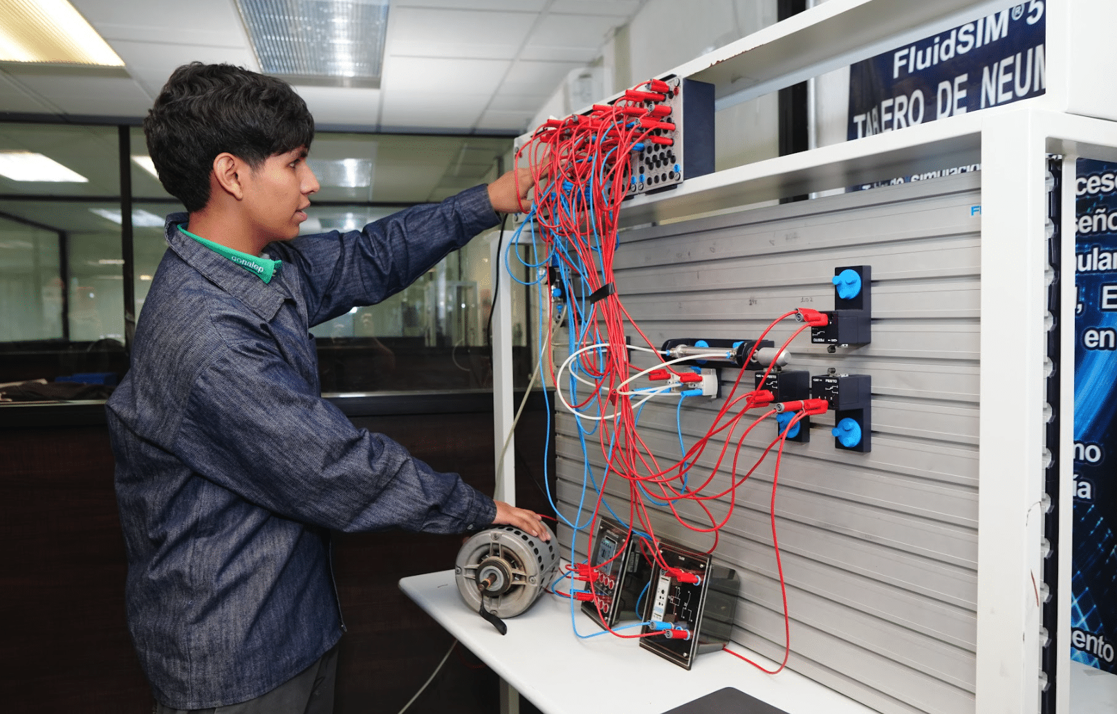 A person in a blue jacket operates a control panel with many red and blue wires in a laboratory setting, adjusting connections on an experimental setup involving electronics and machinery.