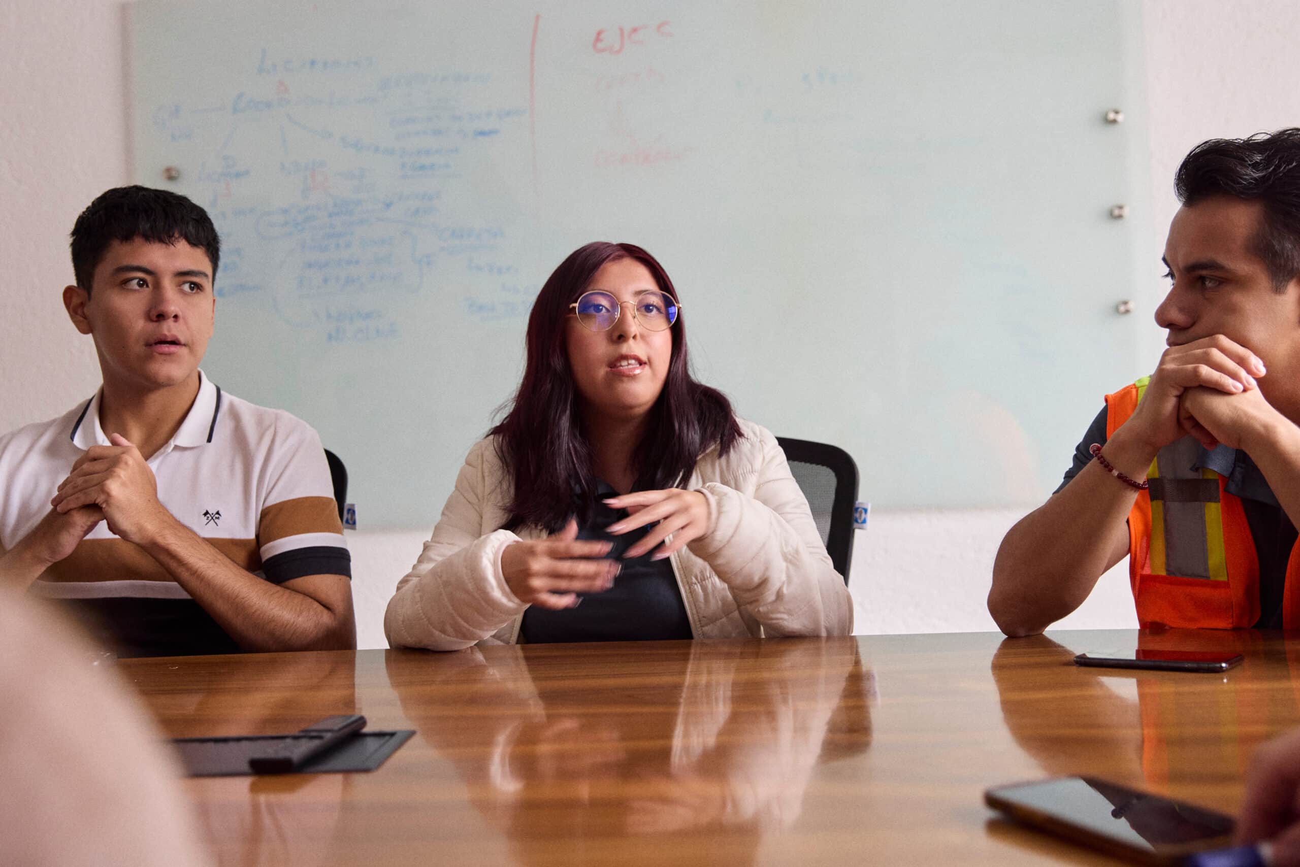 Three people sit at a conference table in discussion. A whiteboard with notes is visible behind them. The person in the center gestures with hands, while the others listen attentively. One person wears a safety vest.
