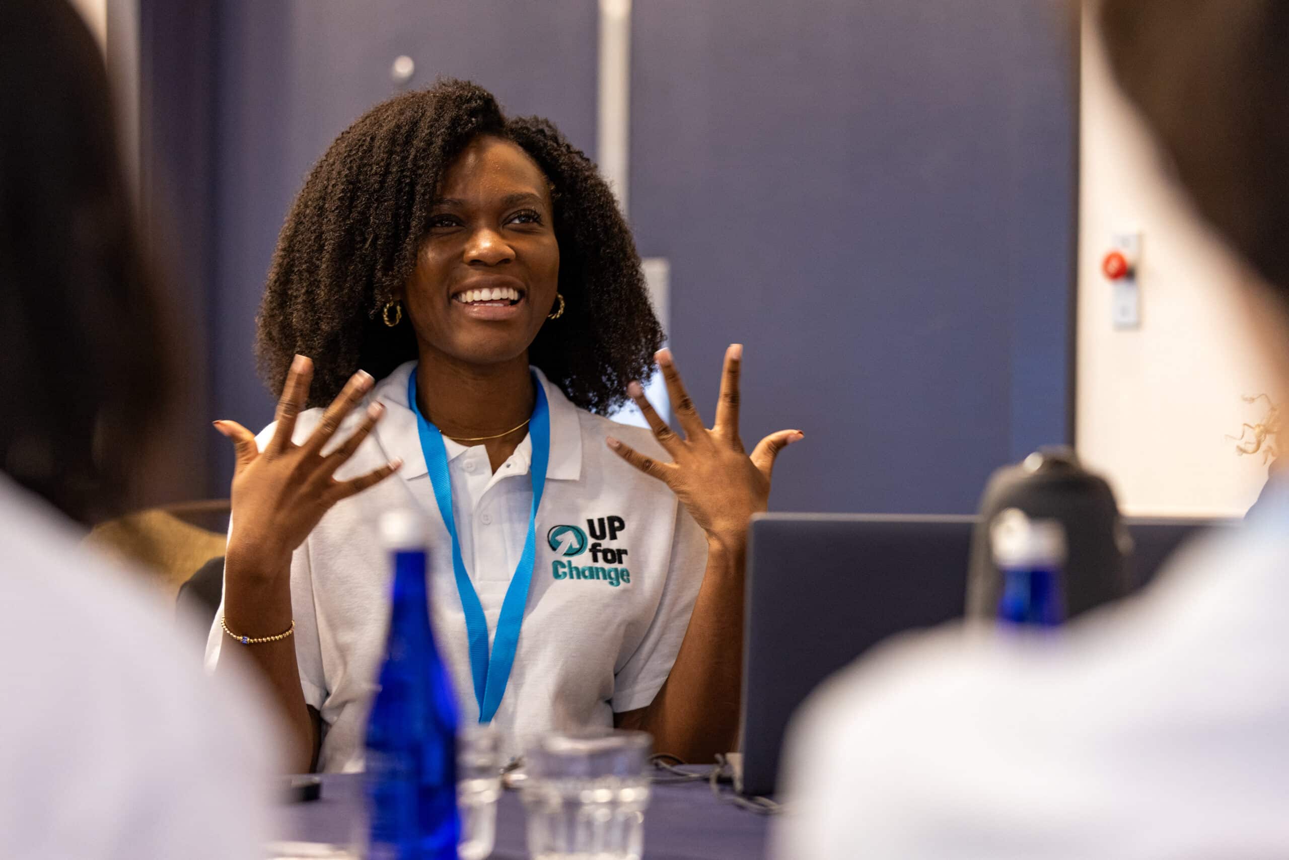 A woman with curly hair wearing a white UP For Change polo shirt and blue lanyard sits at a table, smiling and gesturing with her hands. There are water bottles and glasses on the table.