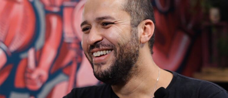 A man with a short beard and buzz cut smiles while sitting indoors, wearing a black shirt and a microphone on his collar. The background features a colorful mural with abstract shapes in red and pink tones.