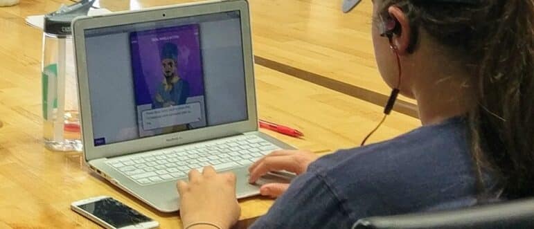 A person wearing headphones uses a laptop at a wooden table; a water bottle, red pen, and smartphone are nearby. The laptop screen displays a digital character and on-screen text.