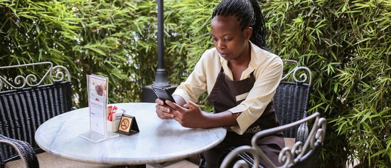 A woman in a work uniform sits at a round outdoor table, looking at her phone. The table has a menu, a napkin holder, and a small no smoking sign. Green plants are in the background.