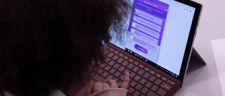 A person with curly hair uses a laptop, interacting with a survey or quiz displayed on the screen. Their finger is touching the keyboard, and a white piece of paper lies on the desk nearby.