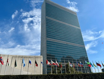 A tall modern glass building with many country flags in front, set against a blue sky with scattered clouds.