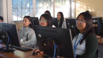 A group of young women sit at desks using desktop computers in a classroom with large windows and natural light.