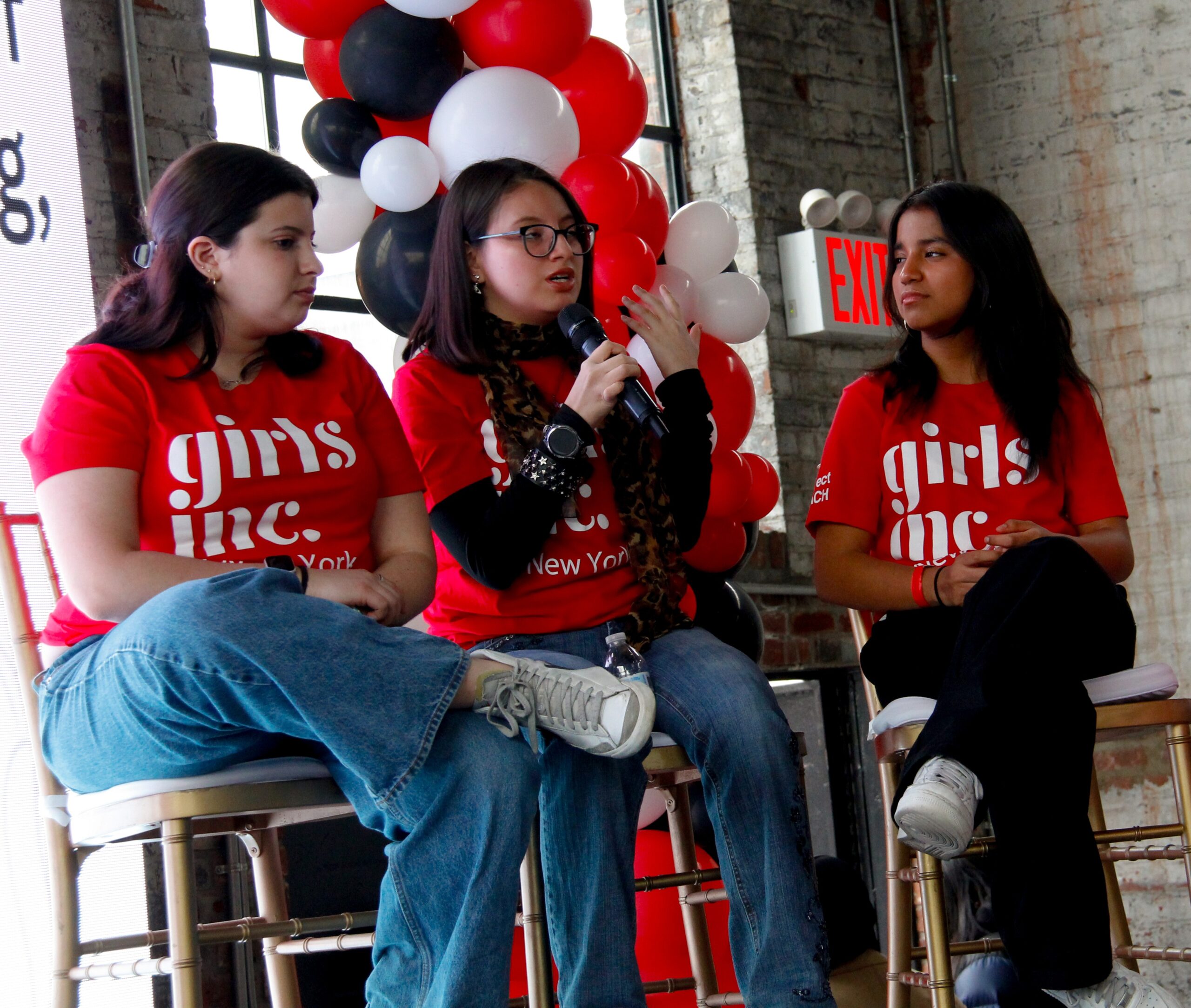 Three young women wearing matching red Girls Inc. New York shirts sit on chairs indoors. One speaks into a microphone while the others listen. Behind them are red, white, and black balloons and a brick wall with windows.