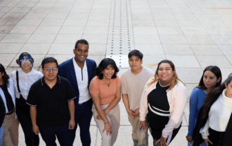 A group of nine people stands outdoors on a tiled surface, posing for a photo and looking up at the camera. They are dressed in casual and business-casual clothing. The background is an open paved area.