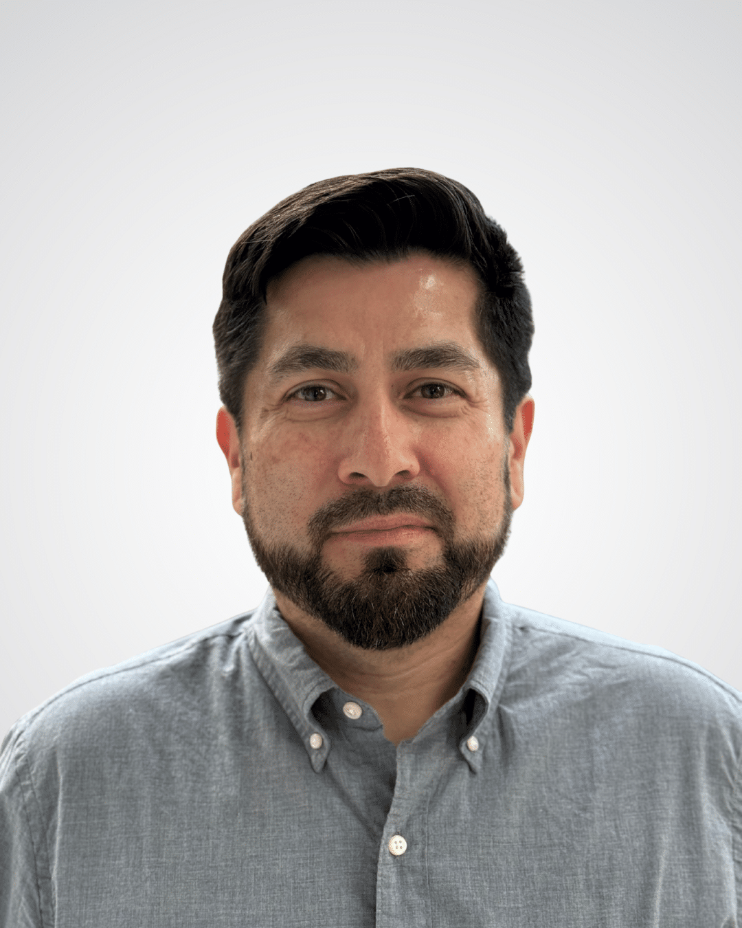 A man with dark hair and a beard, wearing a light gray button-up shirt, looks directly at the camera against a plain light gray background.